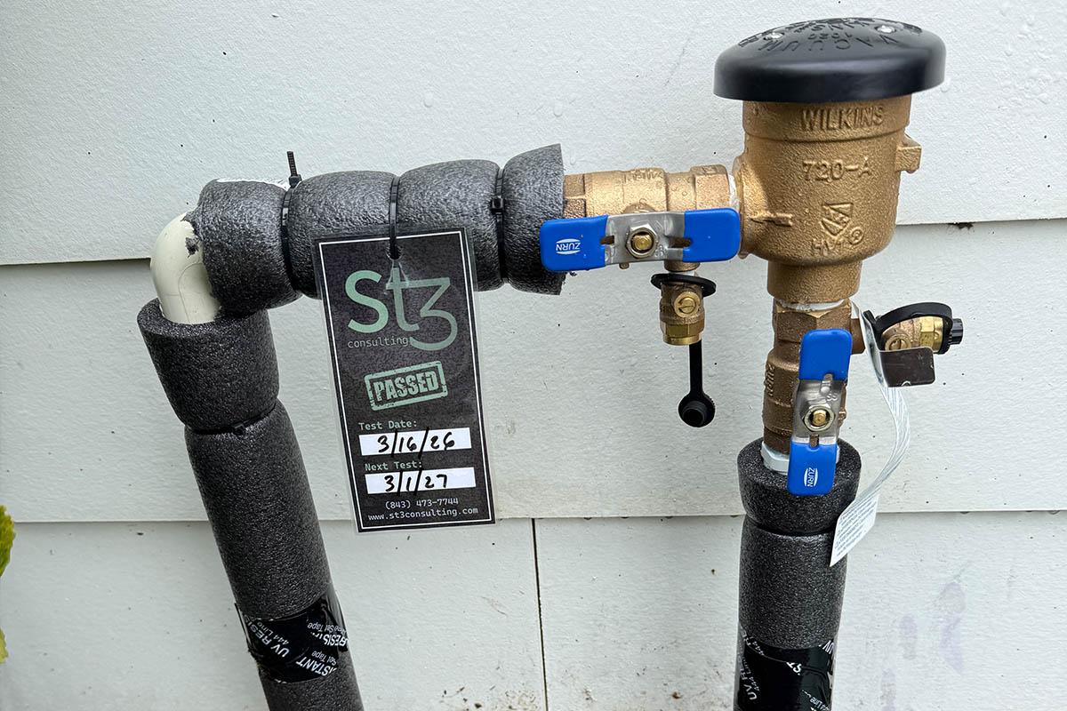 A residential irrigation backflow assembly with an ST3 Consulting “passed” test tag showing the test and next-test dates