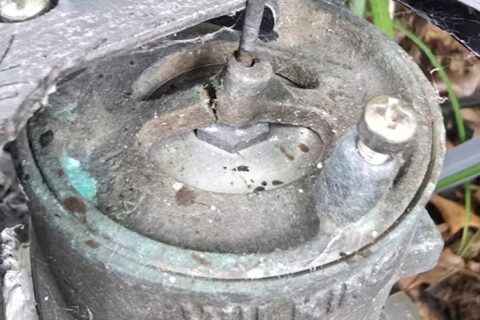 Corroded outdoor electrical junction box with damaged cover, surrounded by grass and fallen leaves.